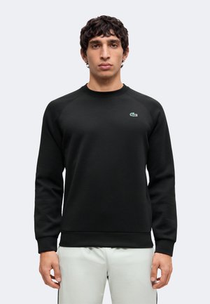 Jeune homme aux cheveux bouclés foncés portant un sweat-shirt noir à manches longues avec un petit logo crocodile vert sur la poitrine.