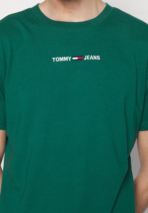Zielona koszulka z krótkim rękawem z haftowanym na środku klatki piersiowej napisem "TOMMY JEANS" oraz małą czerwoną, białą i niebieską flagą.