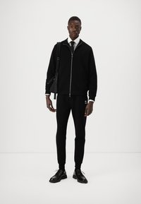 Schwarze Zip-up-Trainingsjacke mit hohem Kragen, kombiniert mit schwarzen Slim-Fit-Hosen. Schwarze Dressschuhe runden das Outfit ab. Einfaches und schickes Design.
