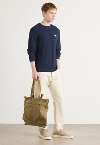 Marineblauwe longsleeve, crèmekleurige broek, beige sneakers; houdt een olijfgroene tote bag vast met twee bovenhandvatten en drukknoopsluitingen.