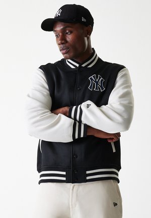 Giacca varsity in bianco e nero con logo NY, colletto e polsini a righe a coste, chiusura con bottoni a pressione e maniche a contrasto.