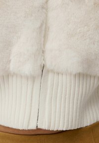 Kratší kardigan z umělé kožešiny v krémové barvě, s žebrovanou pletenou texturou na lemu, se středním zadním švem a jemným leskem díky detailu na zipu.