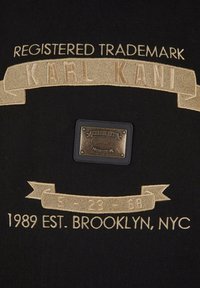 Black fabric featuring gold embroidery of "REGISTERED TRADEMARK" and "KARL KANI." A rectangular black label with metallic accents is included.