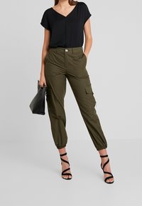 Top negro de manga corta con escote en V combinado con pantalones cargo verde oliva que tienen puños elásticos y múltiples bolsillos. Bolso negro y tacones con tiras.