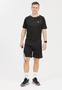 Musta urheilupaita, jossa on hienovarainen kuvio, yhdistetty mustiin shortseihin. Asusteena mustat ja vaaleanpunaiset urheilukengät sekä valkoiset sukat. Pitelee mailaa.