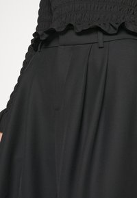 Gros plan sur un pantalon noir plissé taille haute associé à un haut noir texturé smocké avec un ourlet à volants.