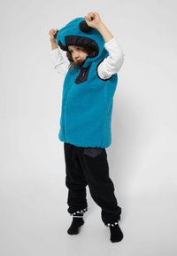 Türkisfarbene Fleece-Weste mit schwarzen Akzenten, ausgestattet mit einer Kapuze mit Ohren. Kombiniert mit schwarzen Fleece-Hosen, die beide eine texturierte Oberfläche haben.