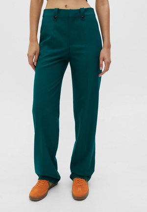 Woman wearing high-waist teal trousers with button details and orange sneakers, standing against a plain gray background.