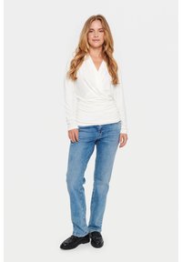 White long-sleeve wrap top with gathered detail, paired with light blue straight-leg jeans and black shoes, against a plain background.