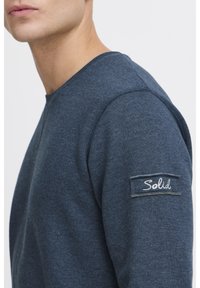 Sudadera azul marino hecha de un tejido suave, con cuello redondo y un parche en la manga izquierda con la palabra "Solid" bordada en blanco.