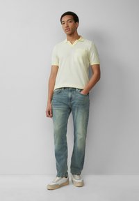 Jasnożółta koszulka polo z dekoltem w serek i kieszonką na piersi, w połączeniu z wyblakłymi niebieskimi dżinsami oraz białymi sneakersami z szarymi akcentami i beżową podeszwą.