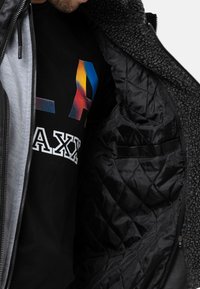 Schwarze Jacke mit einem strukturierten, flauschigen Innenfutter, die eine glatte Außenseite und ein gestepptes schwarzes Innenfutter aufweist. Ein grafisches T-Shirt ist teilweise sichtbar darunter.