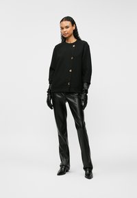 Γυναίκα με μαύρο κουμπωτό πουλόβερ, μαύρα δερμάτινα παντελόνια, μαύρα δερμάτινα γάντια και μαύρα μυτερά παπούτσια, standing σε λευκό φόντο.