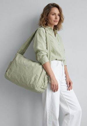 Vrouw draagt lichtgroen overhemd met knopen en witte broek, houdt grote bijpassende lichtgroene schoudertas vast tegen een grijze achtergrond.