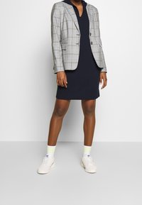 Blazer revisado en gris y azul sobre un vestido con capucha azul marino. Zapatillas blancas con calcetines de rayas en verde claro y amarillo. Atuendo simple y casual.