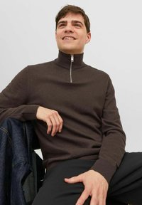 Pull en maille marron à col montant avec fermeture éclair. Il possède des manches longues, des poignets côtelés et une texture lisse. Porté avec un pantalon noir.