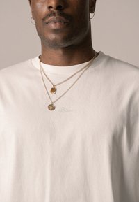 Weißes Baumwoll-T-Shirt mit "Blkts" in hellgrün gestickt. Es hat drei übereinanderliegende goldene Halsketten – eine mit einem rechteckigen Anhänger und eine andere mit einem runden Charm.