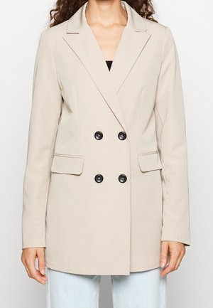 Mujer con blazer beige cruzado con botones negros y bolsillos de solapa, combinado con vaqueros azul claro sobre un fondo liso.
