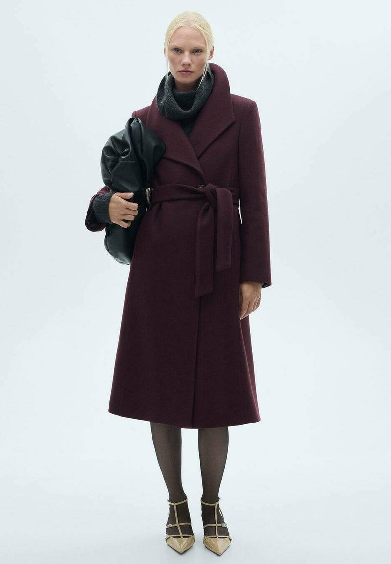 Vinharskåls uldomslagsjakke med bælte, stor krave, båret over en mørk sweater, parret med sorte bukser og sko med guldaccent.