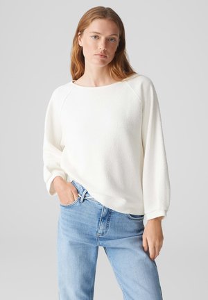 White textured pullover with a wide boat neckline and raglan sleeves. Paired with light blue jeans. The fabric has a tactile surface.