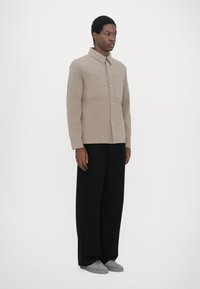 Tan button-up shirt with two chest pockets, paired with wide black pants and gray shoes. Fabric appears textured and casual. Minimalist design.