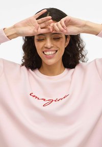 Lyserosa genser med løs passform og rund hals. Har rød brodert tekst "imagine" på fremsiden. Myk stofftekstur.