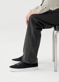 Pantalon en denim noir et baskets en daim foncé avec semelles en caoutchouc blanches, vues de côté en position assise sur une chaise.