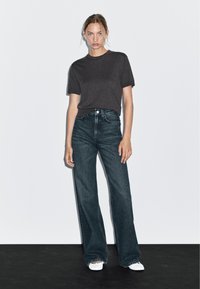 T-shirt grigio scuro a maniche corte abbinato a jeans in denim scuro a vita alta e a gamba larga. Modella con scarpe bianche su uno sfondo semplice.