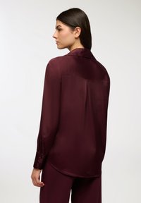Bordowa satynowa koszula o gładkiej fakturze, z długimi rękawami i spiczastym kołnierzykiem; ma plisę z tyłu oraz guziki na mankietach.