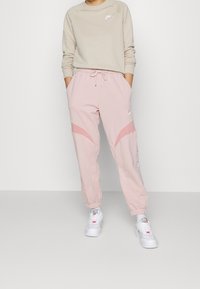 Jasnobeżowa bluza z okrągłym dekoltem i białym logo, w połączeniu z różowymi dresami z zakrzywionymi detalami i ściągaczami, do tego białe trampki.