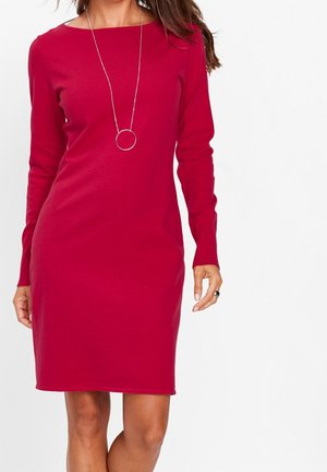Vrouw draagt een lange mouw, knielange rode jurk en een lange zilveren ketting met een cirkelvormige hanger tegen een effen witte achtergrond.