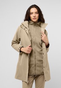 Giacca leggera beige con cappuccio e zip frontale, sovrapposta a una morbida giacca interna in pile beige, dotata di bottoni a pressione.
