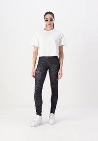 Bela skrajšana majica v kombinaciji s črnimi tajicami z leopardjim vzorcem. Model nosi bele sončna očala in bele teniske. Gladek material, prilegajoč dizajn.