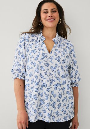 Kobieta uśmiechająca się, ubrana w białą bluzkę z niebieskim kwiatowym wzorem i marszczonymi rękawami, stojąca na jednolitym jasnym tle.