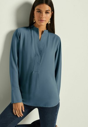 Pelēkzilā krāsā garu piedurknu blūze ar augstu apkakli, ar priekšējo pogu rindu un maigu spīdumu, saskaņota ar tumšiem džinsiem.