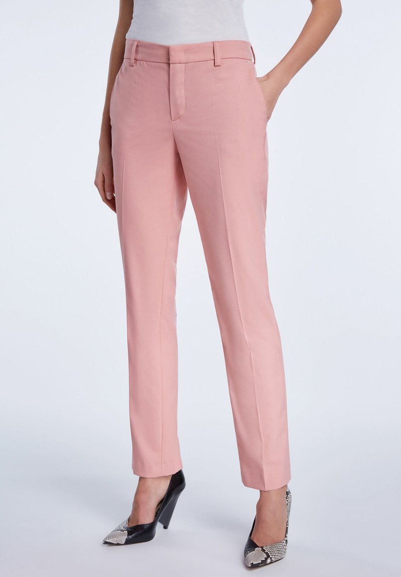 Femme portant un pantalon rose taille haute sur mesure et des talons noirs avec un détail imprimé serpent, debout avec une main dans la poche.