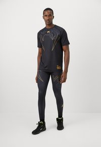 Mann in schwarzem Sport-Shirt und Leggings mit gelbem Schlangendesign sowie schwarzen High-Top-Sneakern, der vor einem weißen Hintergrund steht.