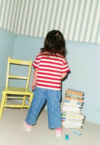 Child wearing a red and white striped shirt, blue denim pants with cherry patterns, and pink shoes, standing beside a yellow chair and book stack.