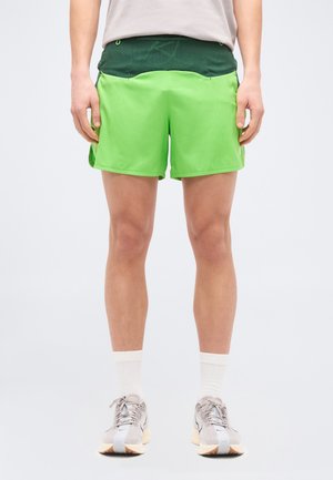 Osoba ubrana w jasnozielone krótkie spodenki sportowe z ciemnozielonym pasem, białe skarpetki i szare buty do biegania stojąca na tle jednolitego tła.