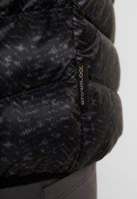 Mjuk svart jacka med texturerad tyg och ett svagt mönster, som visar en etikett med varumärket "Stromlo" i vitt och orange.