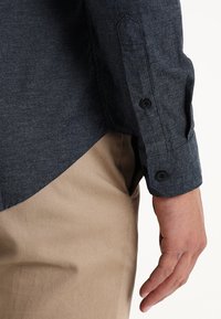 Camisa de manga larga azul oscuro con detalles de puño abotonado, hecha de tela texturizada, combinada con pantalones caqui claros.