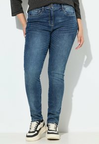 Kobieta w niebieskich wąskich dżinsach i czarno-białych trampkach, stojąca w pobliżu białej ściany z rękami luźno opartymi wzdłuż ciała.