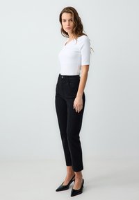 Top bianco a coste con maniche corte, abbinato a jeans neri a vita alta e décolleté nere a punta, mostrato su uno sfondo chiaro.