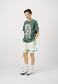 Groen graphic t-shirt met een retro print, gecombineerd met witte gestreepte shorts met groene accenten, zwart-witte sneakers.
