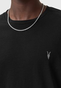 Schwarzes Baumwoll-T-Shirt mit einer kleinen grauen Ziegenkopf-Stickerei auf der linken Brust, kombiniert mit einer verdrehten Silberkette.