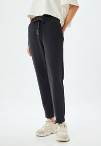 Zwarte, lichtgewicht trainingsbroek met een elastische tailleband en trekkoord. Aangescherpte pijpen. Gecombineerd met lichtgekleurde sportschoenen.
