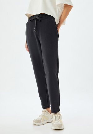 Zwarte, lichtgewicht trainingsbroek met een elastische tailleband en trekkoord. Aangescherpte pijpen. Gecombineerd met lichtgekleurde sportschoenen.