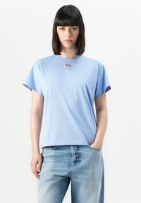 Young woman with straight dark hair wearing light blue t-shirt and faded blue jeans, standing with one hand in pocket against white background.