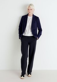 Blazer blu navy su una t-shirt bianca, abbinato a pantaloni neri e sneakers nere con suole bianche. Tessuto morbido con design minimalista.