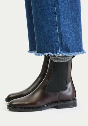 Stivaletti Chelsea in pelle marrone scuro indossati con calzini grigi e jeans blu con orlo sfrangiato su uno sfondo chiaro e semplice.
